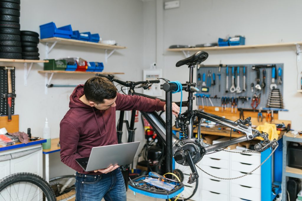 The bike mechanic is manipulating and repairing an electric mountain bike while holding a laptop